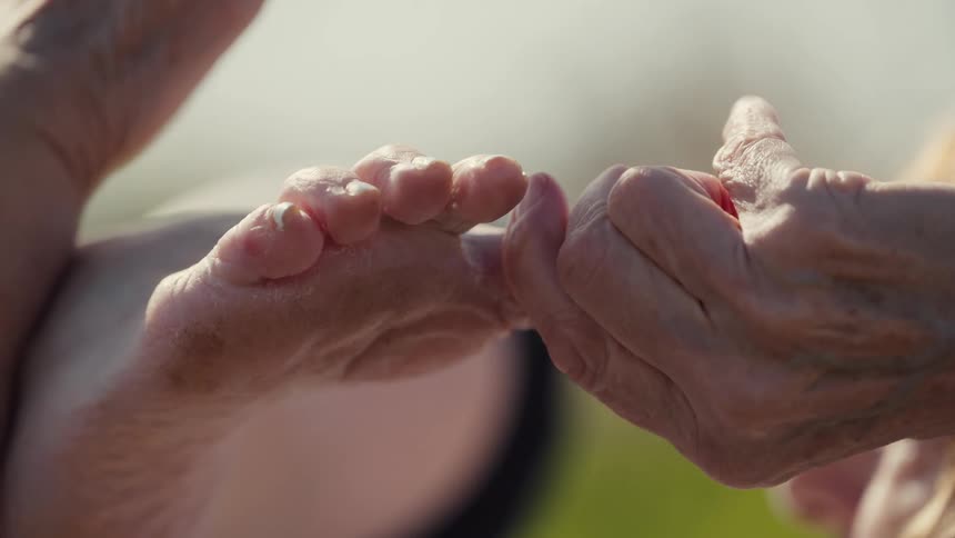 A close up shot where a elderly woman touches her feet with her hand with a blurry background in an outdoor setting