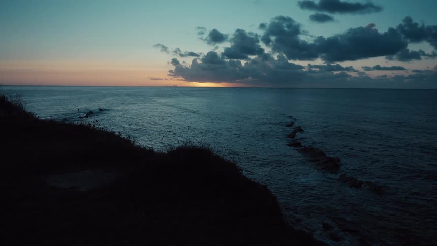 The sun sets on the horizon over a dark calm ocean under a cloudy skya silhouette of a rocky cliffside overlooks the water as the last light fades