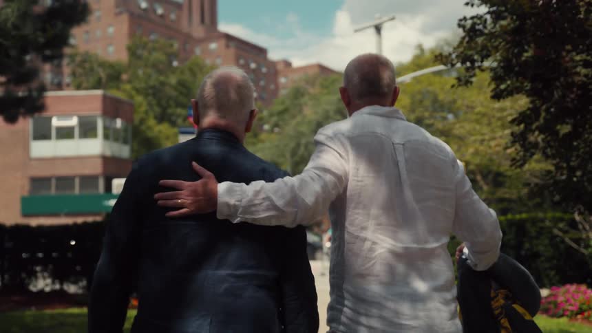 A view from behind shows two older men walking along a city sidewalk as one man places his hand on the shoulder of the other