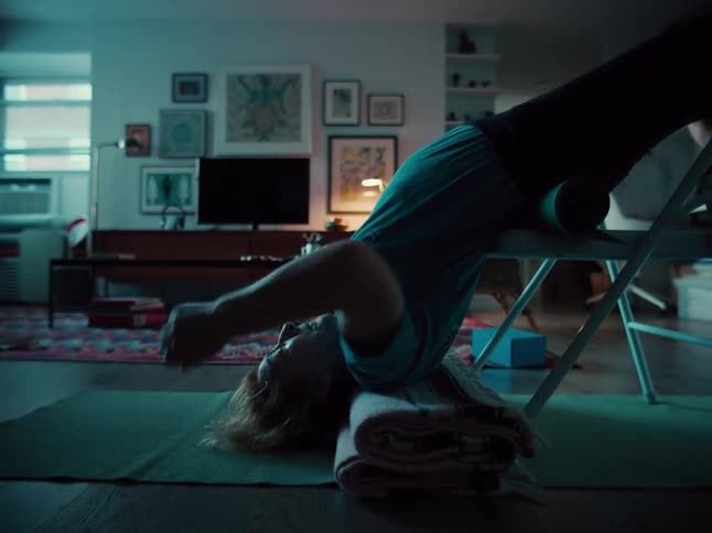 A person performs a restorative yoga pose by arching backward over a metal chair with their head resting on a folded blanketthe scene takes place in a dimly lit cozy living room filled with art frames and a colorful rug