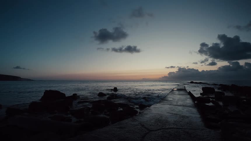 Waves crash against the boulders near a concrete waterfront path as red tints the horizon