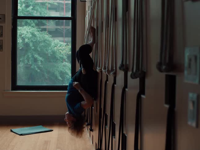 A person is performing an inversion exercise using a yoga wall rope systemthey start by gripping the upper ropes while seated or crouched against the wall then lean back and extend their legs upward to hook their feet into the higher rope loops by th
