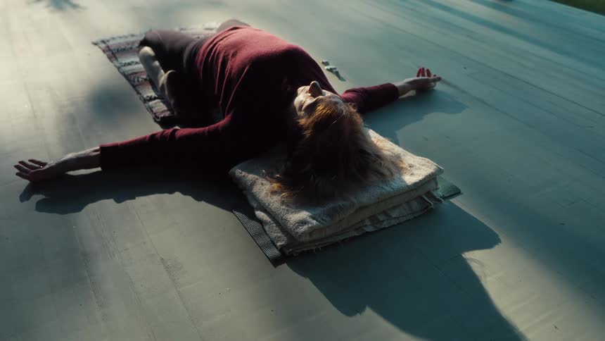 A woman lies on her back on a mat initially extending her arms high above her head before slowly sweeping them down to her sides she remains still in a relaxed meditative pose as sunlight casts long shadows across the floor around her