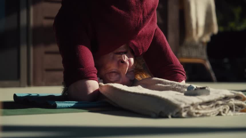 An older person is shown upside down in a yoga pose resting their head on a folded towel atop a blue matthey are wearing a maroon sweater and appear to be focusing intensely on their balance and breathing