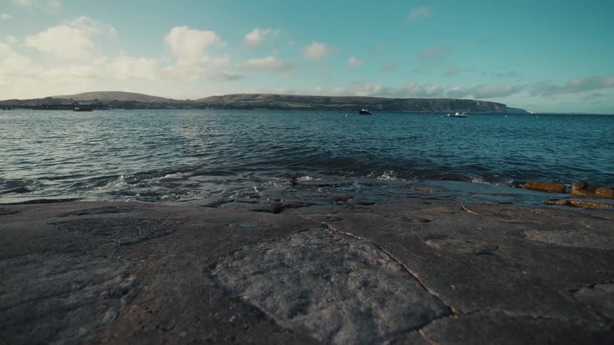 Gently lapping waves wash over a weathered stone pier in a calm coastal bay the scene captures a peaceful morning with distant hills and boats under a bright cloudscattered sky