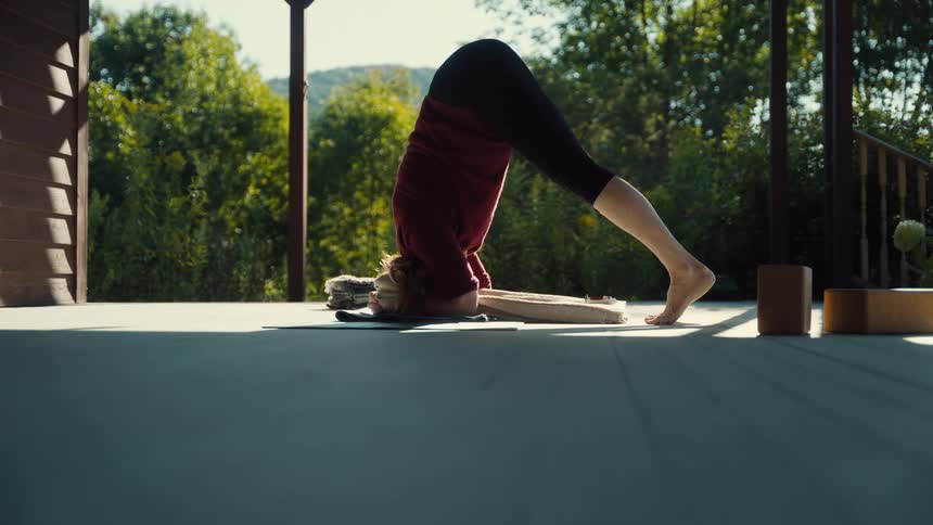 Person performs a headstand with one leg extended on a porch overlooking greenery