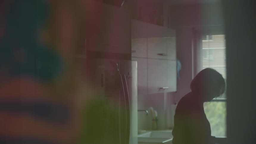 In a dimly lit kitchen a woman is seen in silhouette as she works near a sink and window the shot is captured through a blurred foreground creating a soft and intimate atmosphere as she reaches for something on a shelf