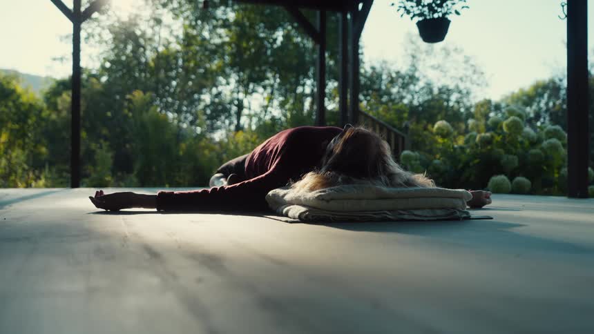 A woman lies peacefully on an outdoor wooden deck resting her head on a soft cushion during a quiet morningsunlight filters through the surrounding lush trees and flowers creating a serene and meditative atmosphere