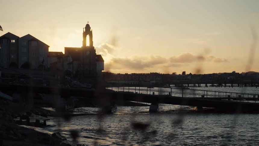 A silhouetted clock tower overlooks a calm coastal town during sunset