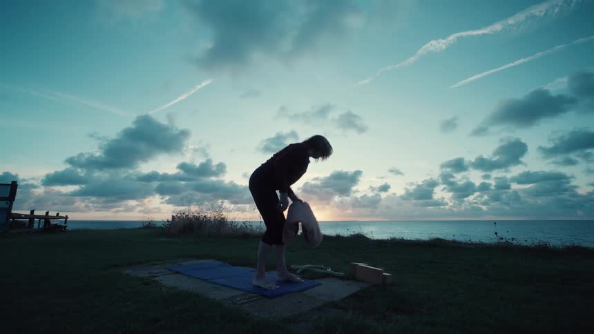 A woman laying a yoga mat on the ground near the coastline in the evening