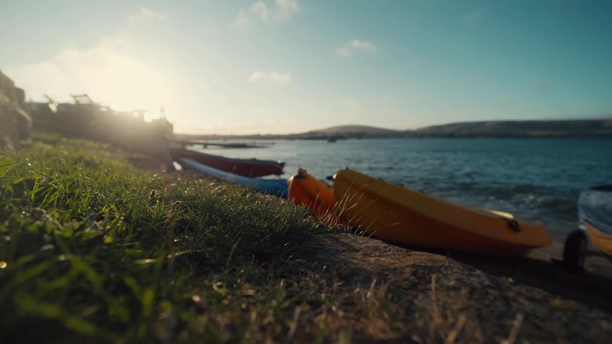 Kayaks rest on a grassy shore by the sea under low sunlight on the horizon
