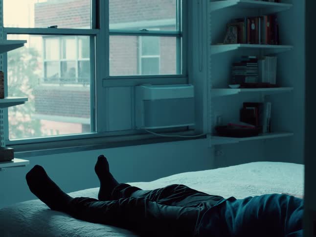 Adult man lying down on a bed in a bedroom indoors next to a window showing trees and a building outside during day time