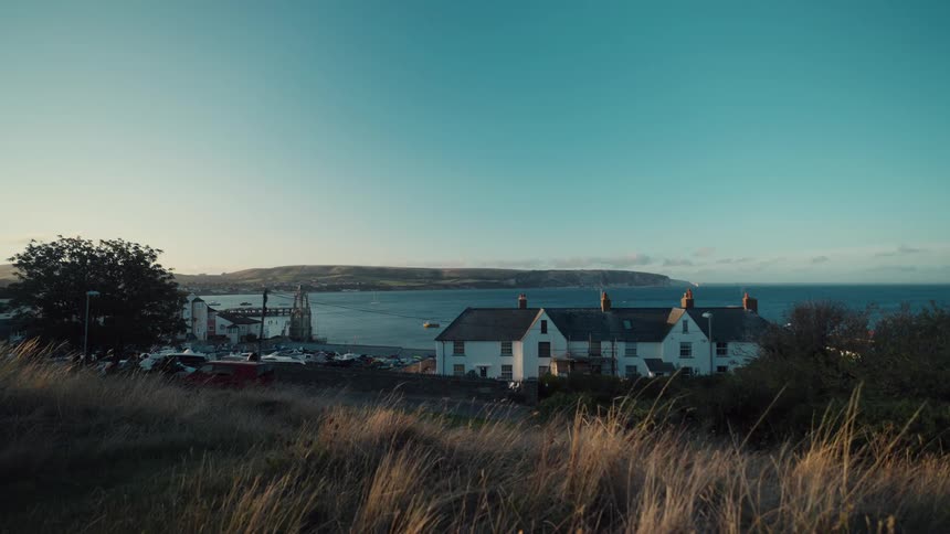 White rural house surrounded by vegetation in front of a street with cars and people next to the ocean outdoors during dusk or dawn
