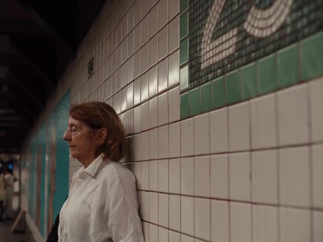 A woman with light brown hair leans against a white tiled wall in a subway stationshe wears a white buttondown shirt and gazes thoughtfully into the distance