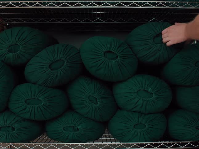 A person wearing a white shirt reaches toward a metal shelf and place one of many dark green bolster cushions stacked together