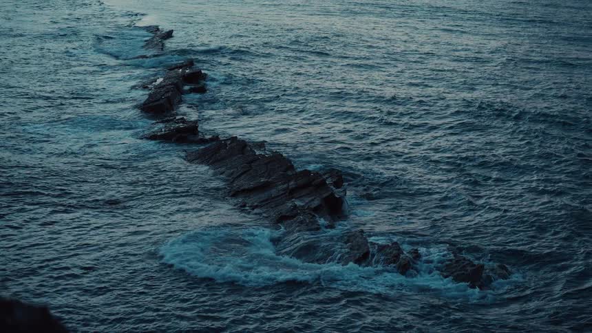 Waves from a dark blue ocean crash against a line of jagged black rocks during the evening