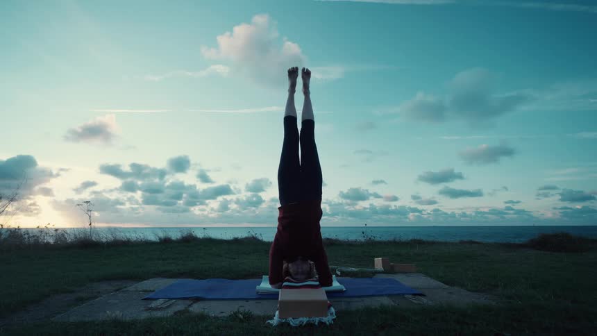 Person performs a headstand on a support in a grassy area near the sea under a cloudy sky