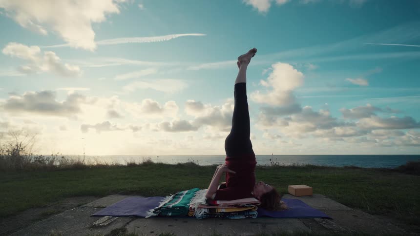 A woman performs a yoga sequence on a cliffside transitioning from a plow pose into a supported shoulder stand set against a coastal sunset she continues the practice by moving her legs into a vertical split while balancing