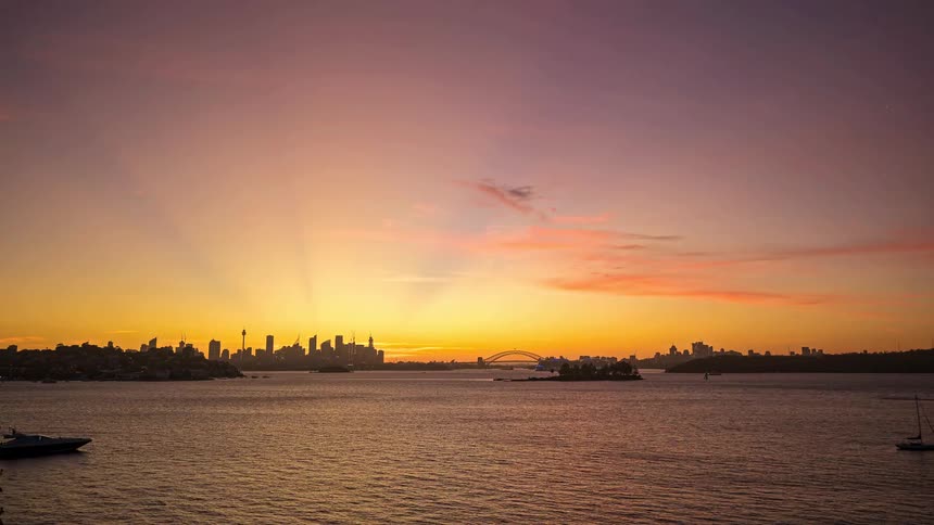 The sun sets behind a city skyline casting an orange glow over the water while a few boats sit anchored in the foreground under a darkening sky