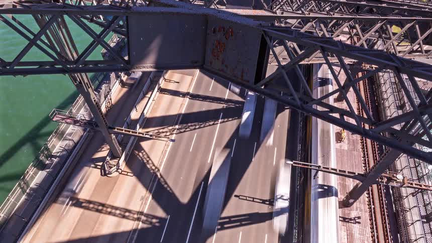 A high angle shot shows cars and trucks driving across the bridge next to empty train tracks while a small boat passes through the green water below the industrial steel trusses and rusted beams of the structure