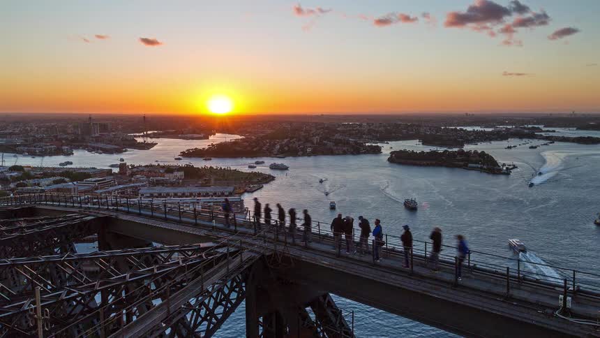 A large group of people crossing a steel bridge as boats pass on the river below with a city skyline and an orange sunset in the distance