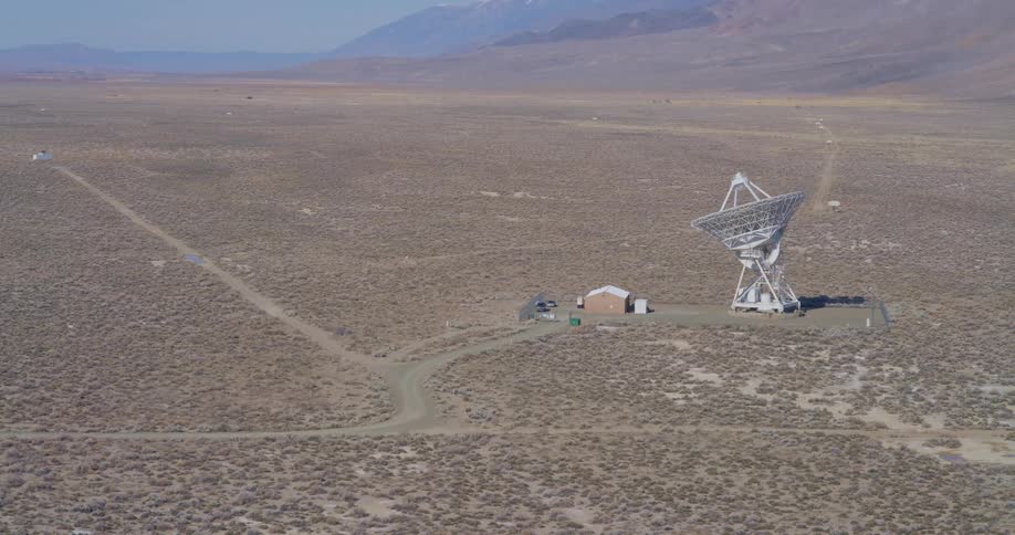 Radio telescope towers over a rural building on a plain bordering the mountains on a foggy day