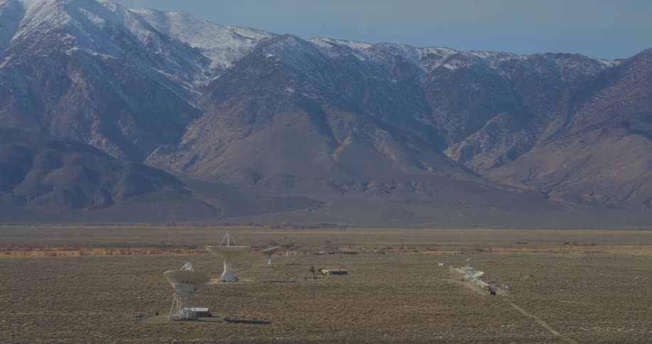 Several large white satellite dishes stand in a flat desert valley with a tall mountain range featuring snow on the peaks in the background