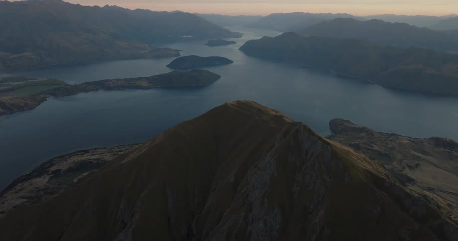 A high angle view shows the peak of a steep mountain overlooking a large blue lake with several islands and smaller mountain ranges under a hazy sky at dawn