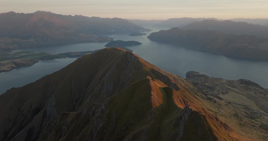 A sharp mountain ridge at dusk revealing a vast lake surrounded by rolling hills and distant peaks under a soft orange sky