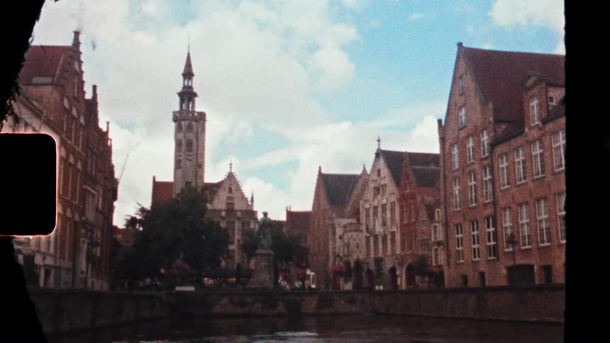 This vintage film footage captures a scenic canal view in bruges belgium featuring traditional brick architecture and the jan van eyck squarethe scene showcases the poortersloge burghers lodge with its distinctive slender spire rising against a p