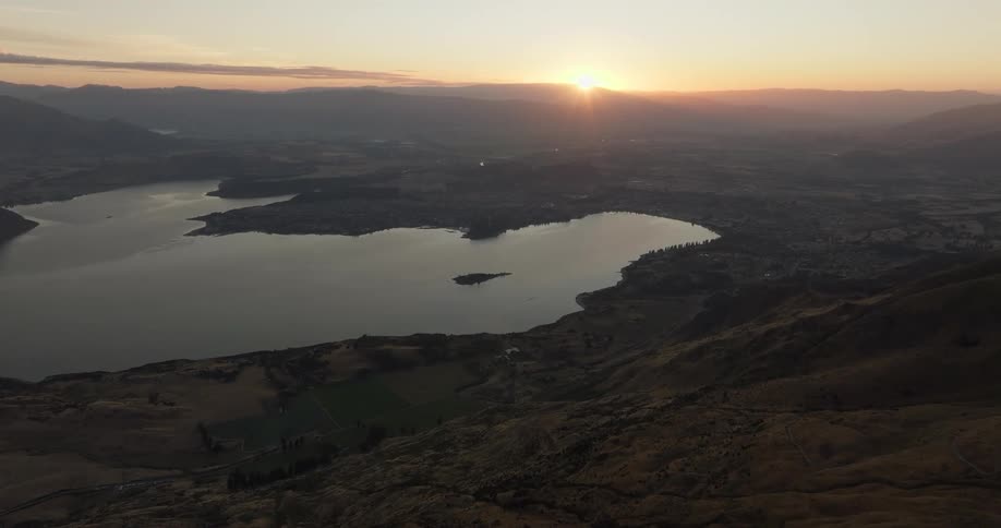An aerial shot reveals a vast landscape where a large calm lake sits nestled among rolling hills and a distant town at sunsetthe golden sun hangs low on the horizon casting a warm glow across the hazy valley and reflecting off the surface of the wat