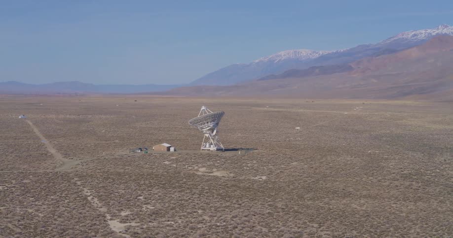 Radio telescope towers over a rural building in a plain located near the mountain on a winter day