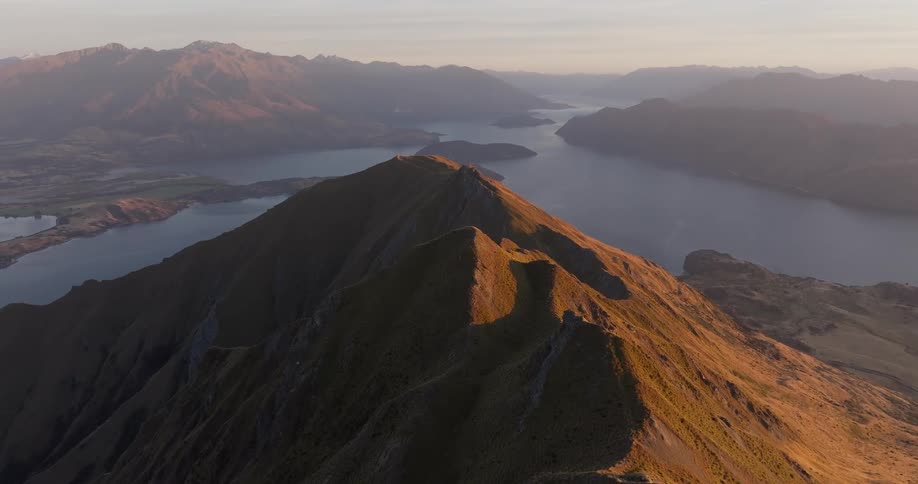 An aerial view reveals the sharp sundrenched ridges of roys peak overlooking the vast expanse of lake wanaka in new zealandthe golden hour light illuminates the winding mountain trail where several hikers can be seen making their way toward the su