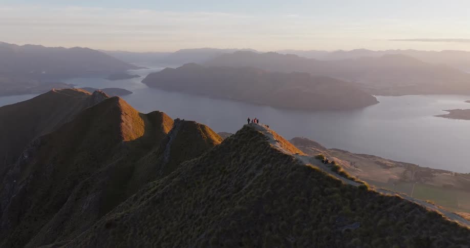 People on a mountain ridge with a lake and mountain ranges in the background under evening light