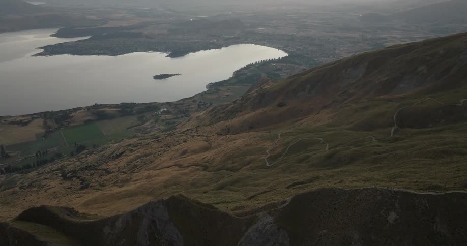 Mountain slopes with visible trails and a lake in the background under soft light
