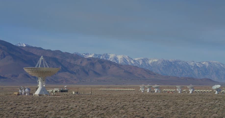 A large white radio telescope stands in a vast desert field with a row of smaller satellite dishes stretching toward the horizon