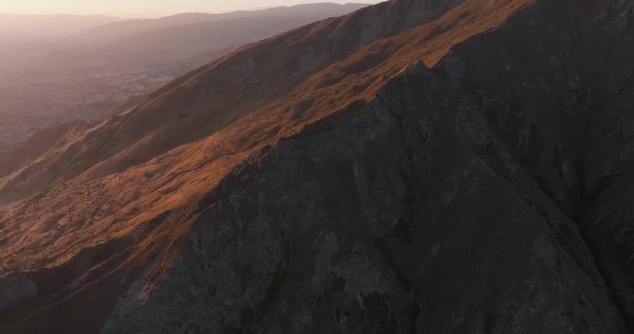 A vast mountain range under the warm glow of a sunset the camera pans across the rugged rocky peaks and grassy slopes capturing the golden light as it hits the side of the mountains