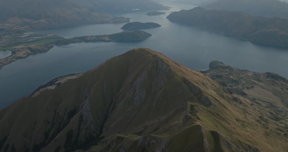 An aerial perspective of a sharp mountain peak overlooking a large winding lake systemas the camera glides forward the rugged ridgeline of the mountain leads the eye toward the deep blue water and distant hills