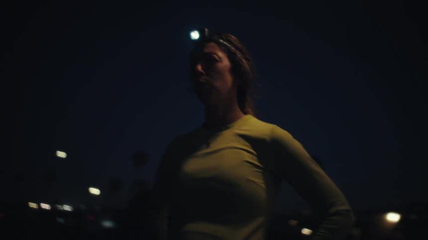 A woman stands outdoors at dusk wearing a yellow longsleeved shirt and a headlampshe rests her hands on her hips while catching her breath and looking out toward the horizon