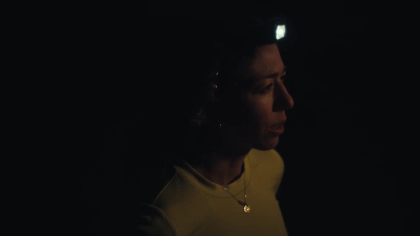 A woman standing in profile against a dark landscape as the first light of dawn begins to glow on the horizon she is wearing a yellow athletic shirt and a lit headlamp looking out into the distance with a focused expression