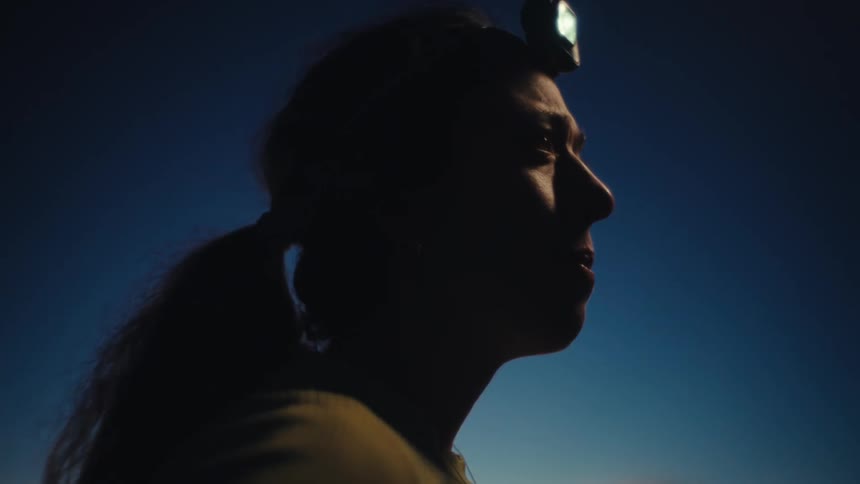 A woman wearing a yellow ribbed shirt and a glowing headlamp looks toward the dark sky before turning her head away