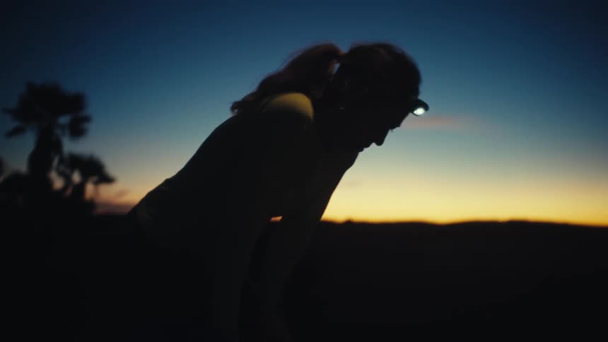 A woman wearing a yellow athletic shirt and a lit headlamp pauses to catch her breath during a twilight run she stands with her hands on her hips and then leans forward against the backdrop of a sunset sky and palm tree silhouettes