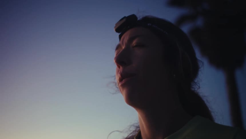 A young woman wearing a headlamp runs through the wilderness then suddenly stops to observe her surroundings as evening gradually turns into night