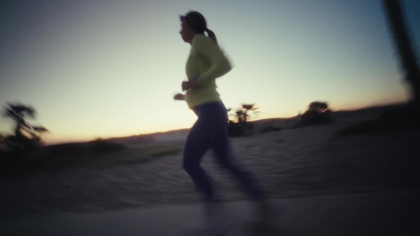 A woman in athletic gear runs along a coastal path during the early morning light her movement is captured in a tracking shot as she passes palm trees under a dim twilight sky