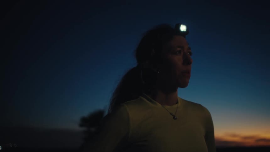 A woman stands in a desert landscape during twilight wearing a headlamp and athletic gear while looking toward the horizonthe camera slowly zooms in on her face as she exhales deeply silhouetted against a fading orange sunset and a dark blue sky
