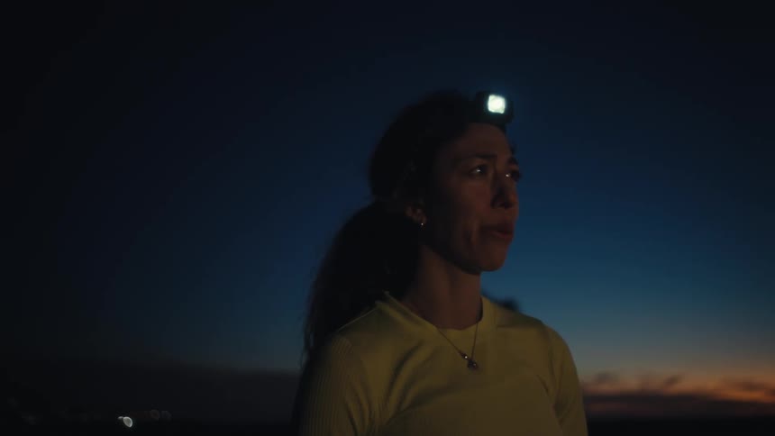 A woman wearing a headlamp stands outside during twilight and breathes deeply while looking into the distance