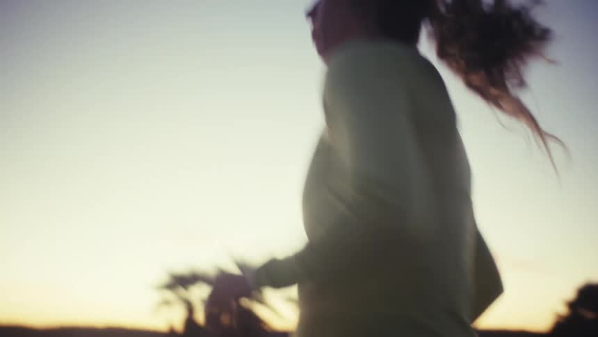 A woman wearing a headlamp and athletic gear runs along a coastal path during a dark sunset the camera captures closeup shots of her stride and form as she moves past silhouetted palm trees