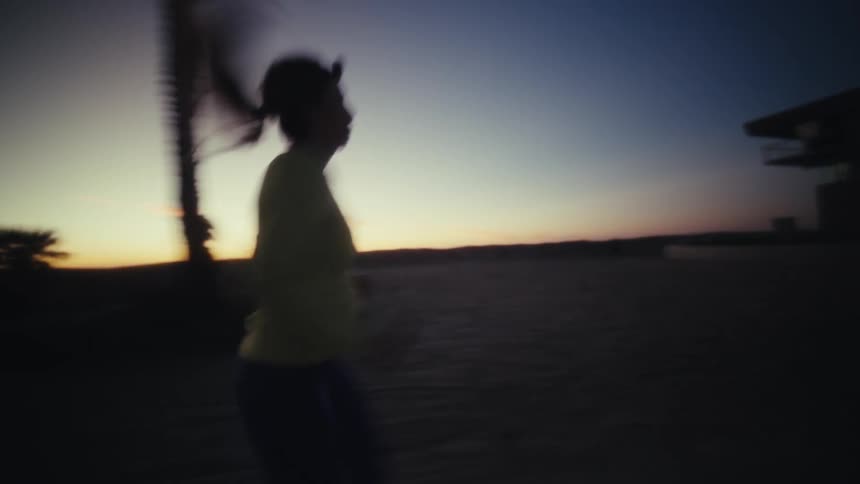 A woman with her hair in a ponytail runs along a path during twilight while wearing a headlampthe camera follows her from the side as she moves past silhouetted palm trees against a fading sunset