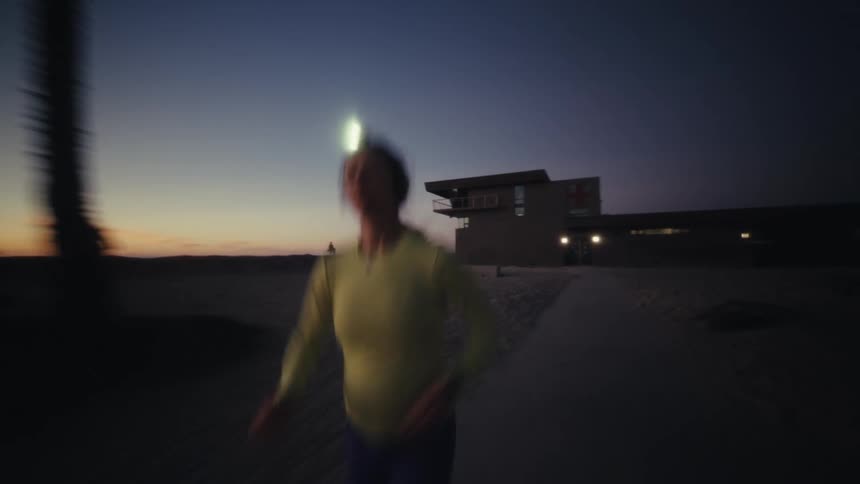A woman wearing a headlamp runs along a beach path during the dim light of dusk the camera follows her closely as she moves past buildings and palm trees silhouetted against a fading orange sky