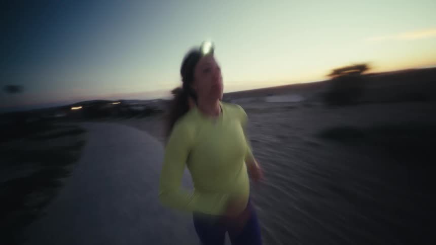 A woman in a yellow longsleeved shirt and blue leggings runs along a coastal path during twilight the camera follows her closely as she passes a modern building near the beach